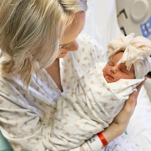 May include: A newborn baby wrapped in a cream-colored blanket with a floral pattern, wearing a matching headband. The baby is held by a person wearing a floral patterned robe. The scene takes place in a hospital setting.