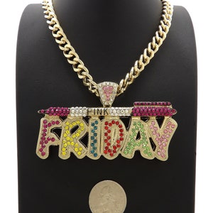 May include: Gold chain necklace with a large pendant that says "PINK FRIDAY" in colourful rhinestones. The pendant is shaped like a banner with a small pink heart at the top.