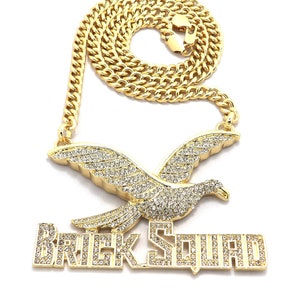 May include: Gold chain necklace with a pendant featuring a bird with wings spread and the words "BRICK SQUAD" set with clear rhinestones.