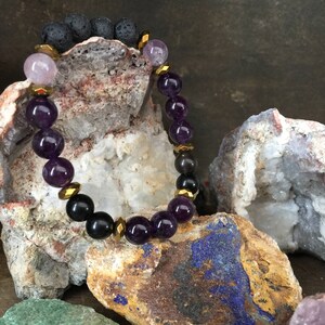 May include: A purple and black beaded bracelet with gold accents. The bracelet is made with amethyst, black lava rock, and hematite beads. The bracelet is displayed on a rock background.