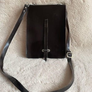 May include: A dark brown leather messenger bag with a long adjustable strap. The bag has a rectangular shape with a flap closure and a buckle detail. The strap is attached to the bag with metal hardware.