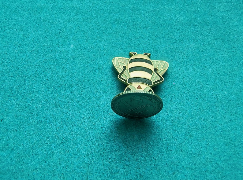 May include: A gold-colored bee figurine standing on a 1974 Soviet 5 kopecks coin. The bee has black stripes and a red stinger.