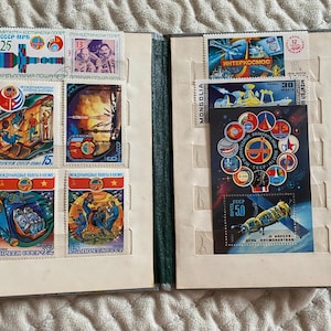 May include: An open stamp album featuring a collection of vintage postage stamps with space exploration themes. The stamps showcase colorful illustrations of rockets, astronauts, and space stations. The album is open, revealing multiple pages filled with stamps.