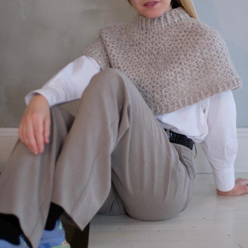 May include: A woman wearing a white shirt and gray knitted cowl neck sweater, sitting with her legs crossed, wearing gray pants.