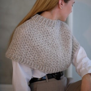 May include: A light gray crocheted cowl with a wide, rounded neckline. The cowl is worn over a white shirt and brown pants.