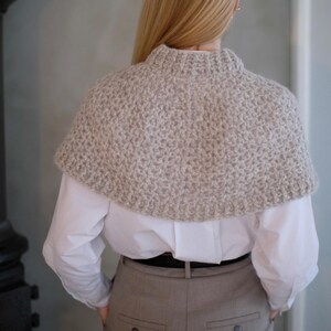 May include: A beige knitted shawl with a high neckline. The shawl is made of a soft, fuzzy yarn and has a simple, elegant design.