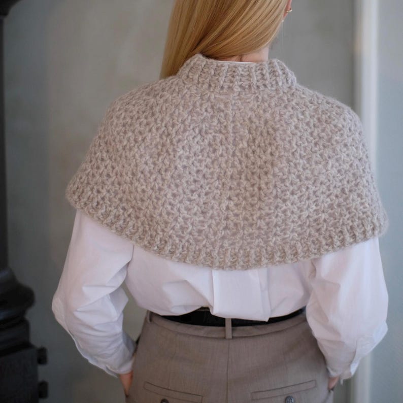 May include: A beige knitted shawl with a high neckline. The shawl is made of a soft, fuzzy yarn and has a simple, elegant design.