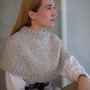 May include: A close-up of a woman wearing a light gray crocheted cowl neck sweater. The sweater is made with a textured stitch and has a loose, comfortable fit.