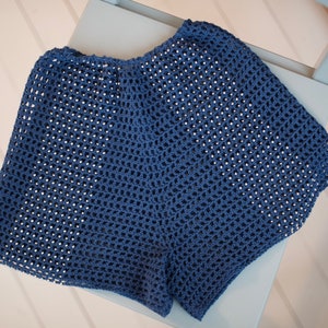May include: A pair of blue crochet shorts with a loose, open weave. The shorts are made of a soft, breathable material and are displayed on a light blue chair. The shorts are a solid color and appear to be handmade.