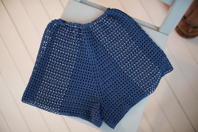May include: A pair of blue crochet shorts with a loose, open weave. The shorts are made of a soft, breathable material and are displayed on a light blue chair. The shorts are a solid color and appear to be handmade.