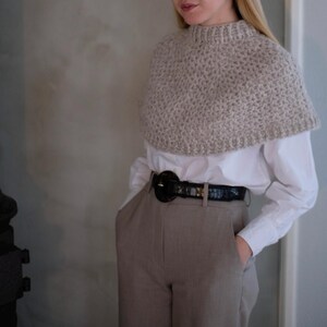 May include: A beige knitted cowl neck sweater with a white button-down shirt underneath. The person is wearing a black belt with a large round buckle and beige pants.