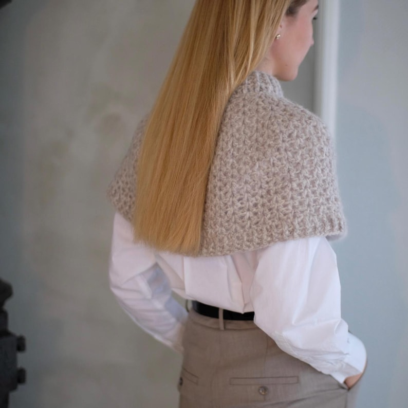 May include: A woman wearing a white shirt and beige pants with a light brown knitted shawl around her neck.