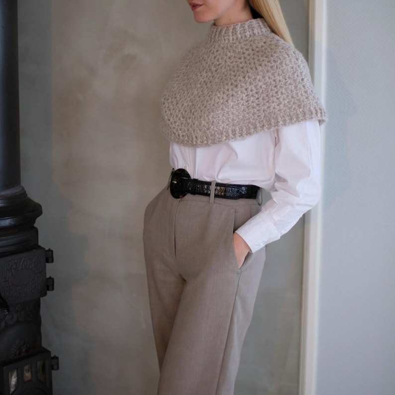 May include: A beige knitted cowl neck sweater with a white button-down shirt and beige pants. The pants have a black belt with a large buckle.