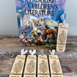May include: A Treasury of Children's Literature book with a colorful illustration and several wooden bookmarks. The bookmarks have floral designs and the text "A New Chapter Begins" and "Thank You For Being A Part Of" with names.