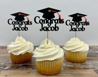 Könnte beinhalten: Drei Cupcakes mit weißem Zuckerguss, jeweils gekrönt mit einer schwarzen Abschlusskappe und den Worten "Congrats Jacob". Die Aufsätze haben rote Quasten und sind auf Holzstäbchen. Die Cupcakes stehen auf einer Holzoberfläche.