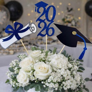 Könnte beinhalten: Ein Abschluss-Tischschmuck mit weißen Rosen und Schleierkraut, verziert mit blauen Glitzer-Ausschnitten zum Thema Abschluss: eine Urkunde, das Jahr 2026 und ein Mortarboard. Ballons und Desserts sind im Hintergrund.