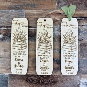 May include: Three wooden bookmarks with a floral and book design. The bookmarks have the text "A New Chapter Begins" and "Thank you for being a part of Emma & Jacob's Story 6.2.26". One bookmark has a green ribbon.