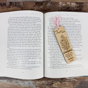 May include: Open book with a wooden bookmark. The bookmark has a floral design and the text "A new chapter begins" and "Thank you for being a part of Amelia's story". A pink ribbon is attached.