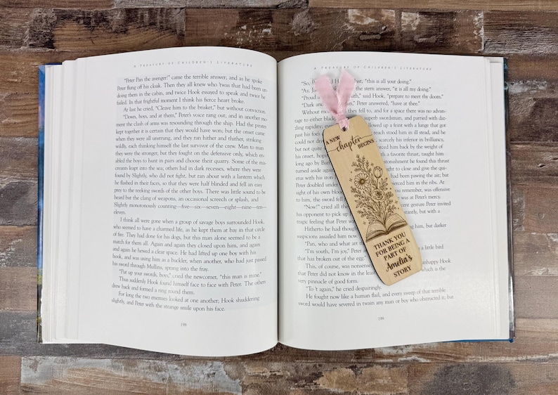 May include: Open book with a wooden bookmark. The bookmark has a floral design and the text "A new chapter begins" and "Thank you for being a part of Amelia's story". A pink ribbon is attached.
