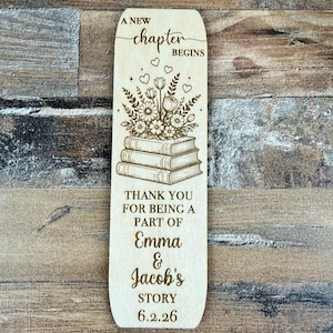 May include: A wooden bookmark with engraved floral and book designs. The text reads "A New Chapter Begins", "Thank You For Being A Part Of Emma & Jacob's Story 6.2.26". The bookmark is a light brown color.