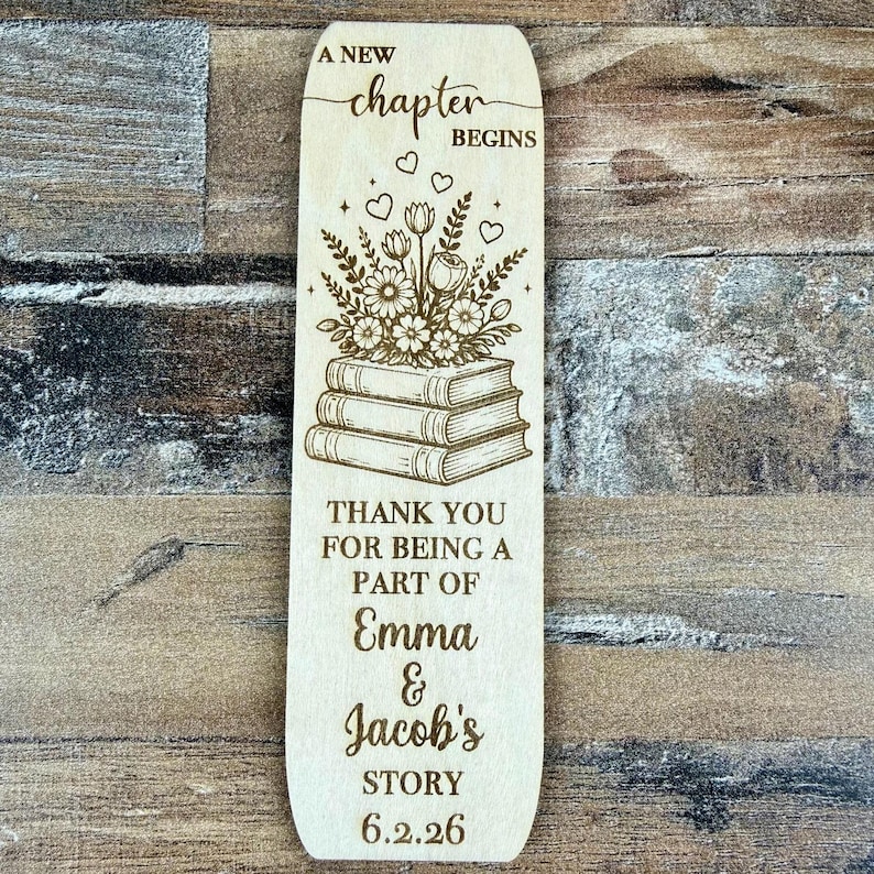 May include: A wooden bookmark with engraved floral and book designs. The text reads "A New Chapter Begins", "Thank You For Being A Part Of Emma & Jacob's Story 6.2.26". The bookmark is a light brown color.