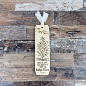 May include: Wooden bookmark with a floral design and the text "A new chapter begins." The bookmark also includes the text "Thank you for being a part of Baby Allen's story." A white ribbon is attached at the top.