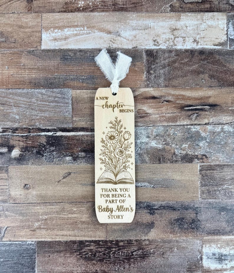 May include: Wooden bookmark with a floral design and the text "A new chapter begins." The bookmark also includes the text "Thank you for being a part of Baby Allen's story." A white ribbon is attached at the top.