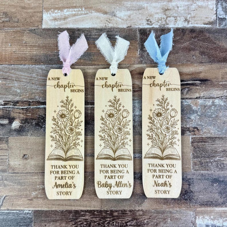 May include: Three wooden bookmarks with floral designs and the text "A new chapter begins." Each bookmark has a different name: Amelia's, Baby Allen's, and Noah's. They have ribbons in pink, white, and blue.