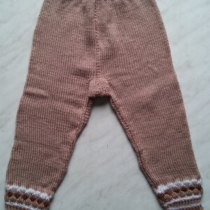 May include: A pair of knitted baby pants in a light brown color with white and brown trim around the ankles.