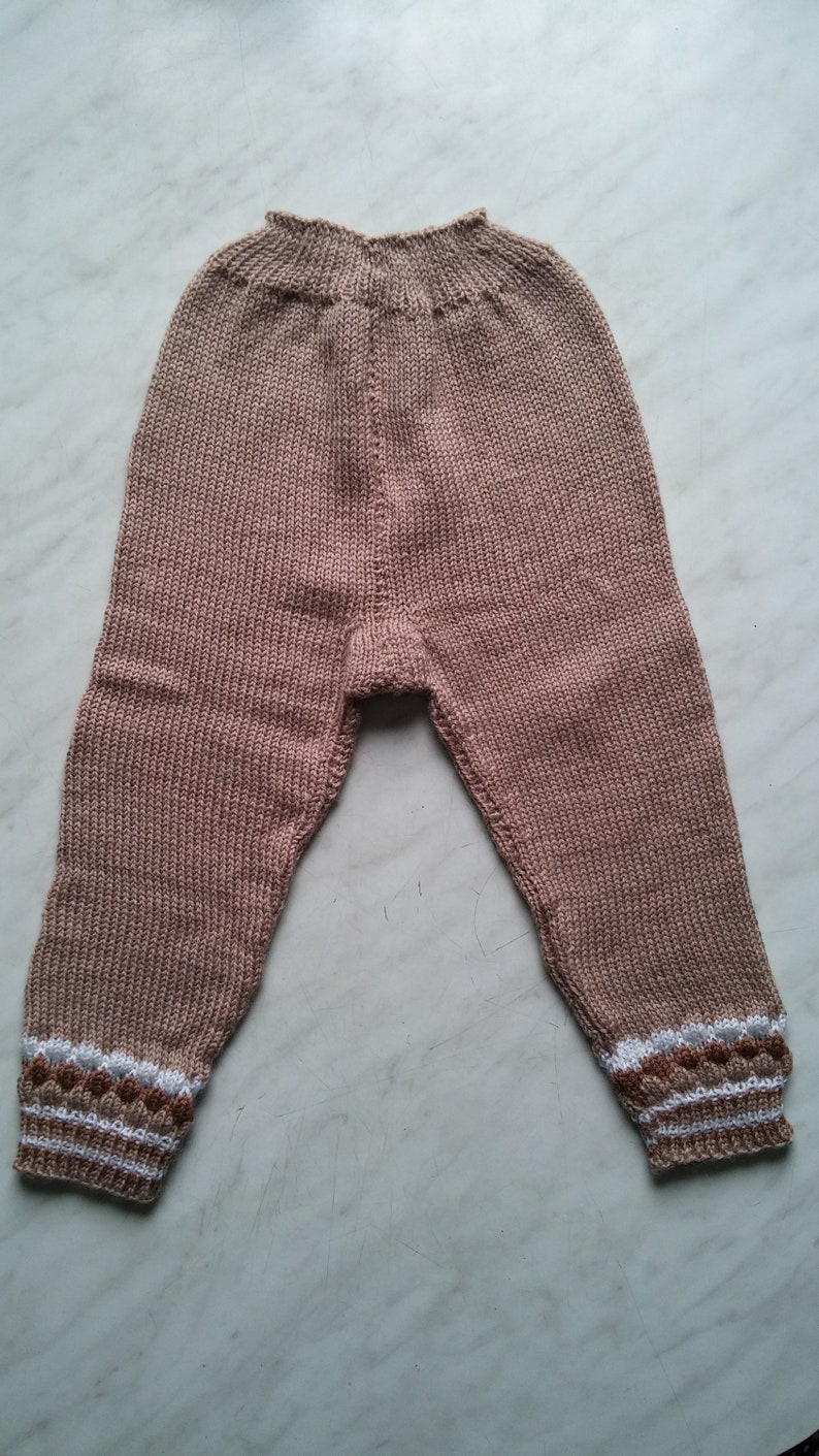 May include: A pair of knitted baby pants in a light brown color with white and brown trim around the ankles.