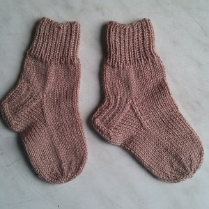 May include: A pair of knitted socks in a light pink color. The socks are made with a simple stitch pattern and have a ribbed cuff.