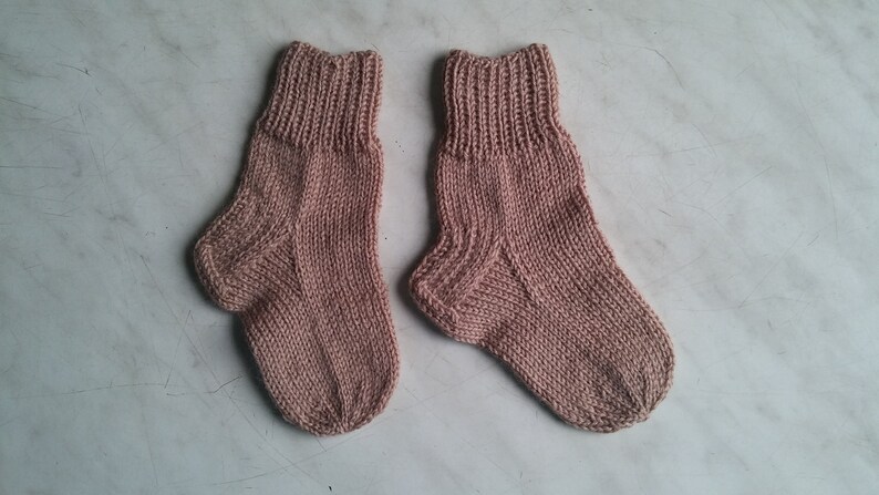 May include: A pair of knitted socks in a light pink color. The socks are made with a simple stitch pattern and have a ribbed cuff.