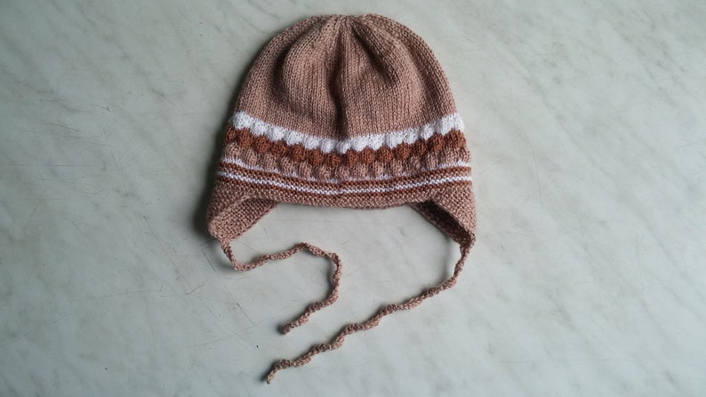 May include: A brown knitted hat with white and brown stripes and ear flaps. The hat has ties that can be tied under the chin.