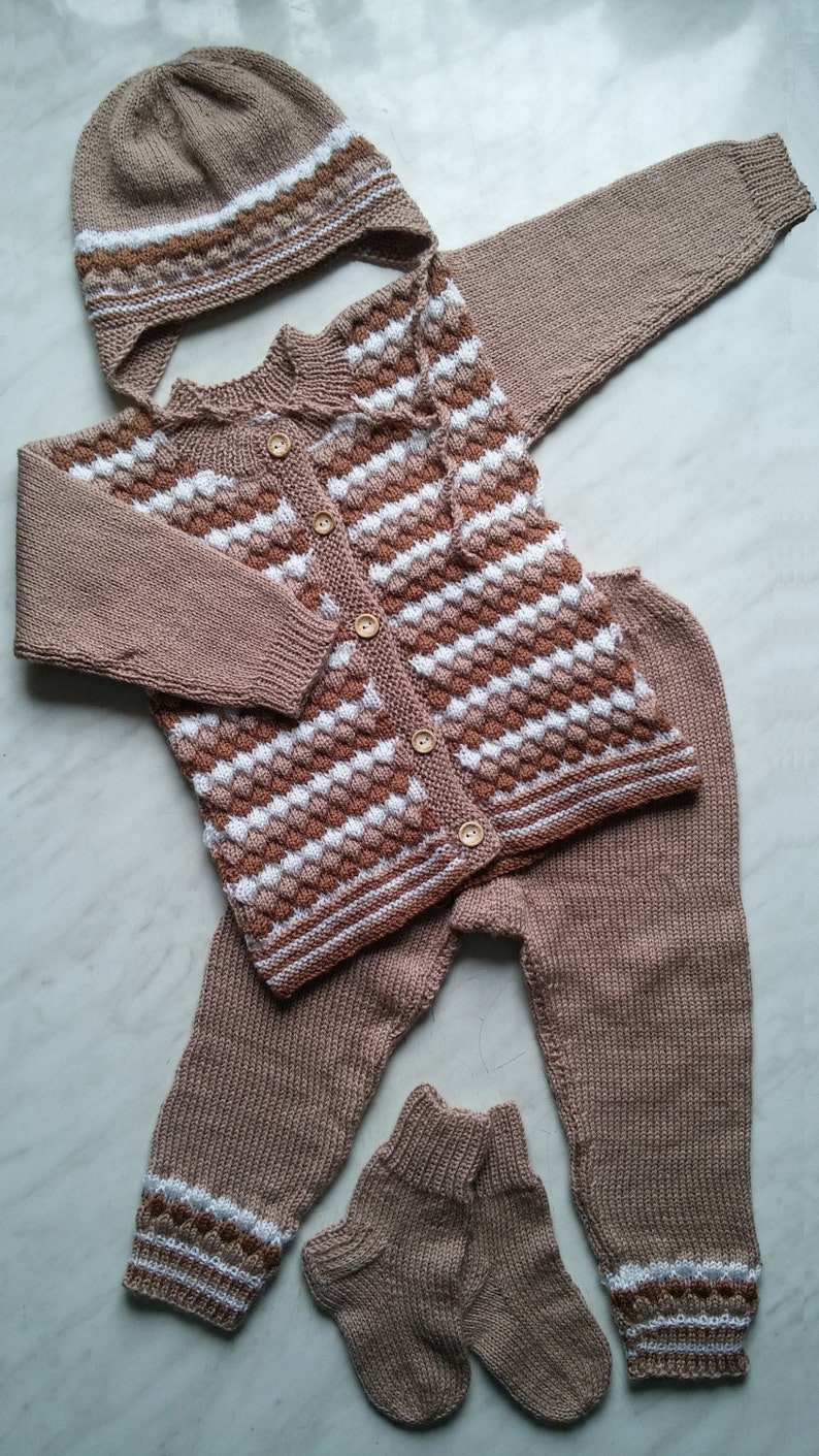 May include: A knitted baby outfit in brown, white, and beige. The outfit includes a hat, sweater, pants, and socks. The sweater has a striped pattern and buttons down the front.