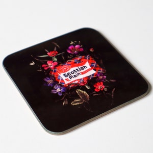 May include: Black coaster featuring a red and white Scottish Plain cigarette pack design surrounded by purple and red flowers.