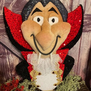 May include: A plush vampire face with a black glittered hair, a large nose, and a smiling mouth. The vampire wears a red sequined cape with black trim and a white ruffled collar. A gold and red decorative piece is below the collar.