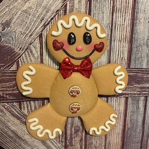 May include: A tan gingerbread person decoration featuring white icing squiggles on the head, arms, and legs. It has black eyes, a pink nose, red glitter heart cheeks, a red glitter bow tie, and two small buttons with gingerbread faces.