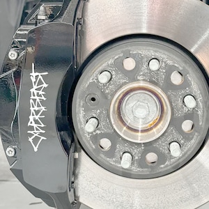 May include: Close-up of a black brake caliper with white graffiti-style lettering that says "SLAUGHTERED". The caliper is attached to a silver brake rotor with a black center hub.