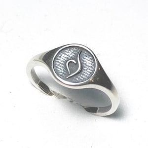 Puede incluir: Un anillo de sello de plata con una parte superior ovalada texturizada que presenta un dise&ntilde;o estilizado.