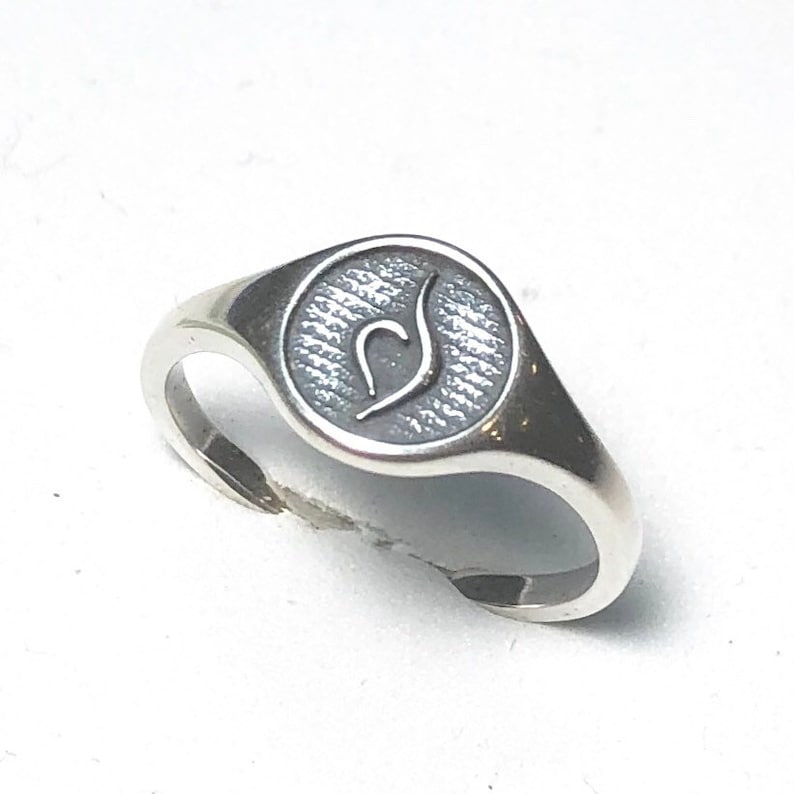 Puede incluir: Un anillo de sello de plata con una parte superior ovalada texturizada que presenta un dise&ntilde;o estilizado.