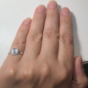 Puede incluir: Un anillo de plata con un peque&ntilde;o s&iacute;mbolo redondo negro en la parte superior. El anillo est&aacute; en el dedo de una persona.