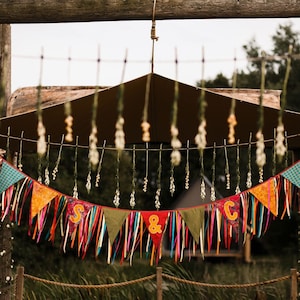 May include: A colorful banner with the letters "S & C" hangs from a wooden structure. The banner features various patterned triangles and long, colorful ribbons. The background includes a brown canopy and greenery, suggesting an outdoor event.