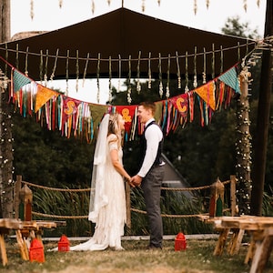 May include: A wedding ceremony setup with a couple holding hands under a decorated canopy. Colorful pennant banners and ribbons hang above. Wooden benches and decorative posts with greenery frame the scene, creating a rustic, outdoor wedding theme.