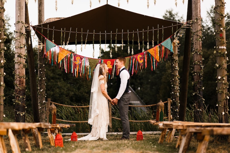 May include: A wedding ceremony setup with a couple holding hands under a decorated canopy. Colorful pennant banners and ribbons hang above. Wooden benches and decorative posts with greenery frame the scene, creating a rustic, outdoor wedding theme.