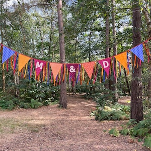 May include: A colorful bunting banner strung between trees in a forest setting. The pennants are in shades of blue, orange, pink, and purple, with the letters "M & D" displayed. The banner is decorated with ribbons.