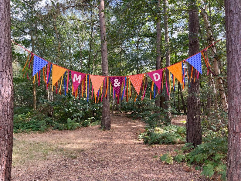 May include: A colorful bunting banner strung between trees in a forest setting. The pennants are in shades of blue, orange, pink, and purple, with the letters "M & D" displayed. The banner is decorated with ribbons.