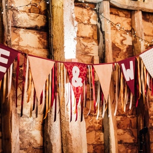 May include: A festive banner with burgundy pennants displaying the letters "E" and "W" and an ampersand symbol. The banner is adorned with ribbons in various colors and hangs against a rustic wooden backdrop, illuminated by string lights.