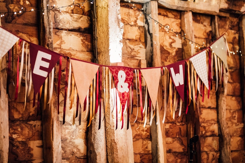 May include: A festive banner with burgundy pennants displaying the letters "E" and "W" and an ampersand symbol. The banner is adorned with ribbons in various colors and hangs against a rustic wooden backdrop, illuminated by string lights.