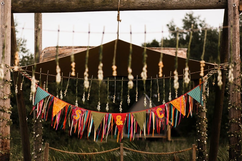May include: A colorful banner with the letters "S & C" hangs from a wooden structure. The banner features various patterned triangles and long, colorful ribbons. The background includes a brown canopy and greenery, suggesting an outdoor event.
