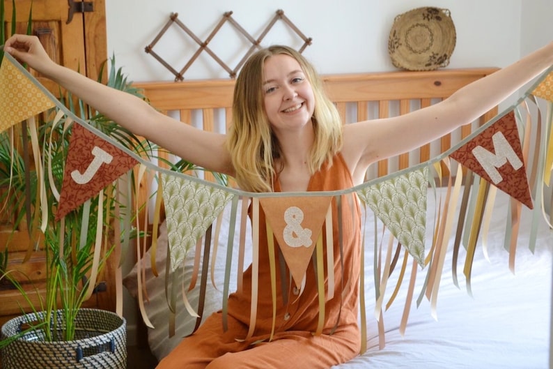 May include: A handmade fabric banner with the letters "J & M" on triangular pennants. The banner features various patterned fabrics and ribbons in shades of brown, beige, and green. Perfect for a wedding or party.